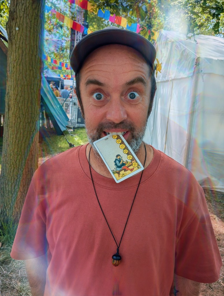 A playful portrait of Beans on Toast at Bearded Theory Festival 2026. Holding the Eight of Pentacles tarot card between his teeth and staring wide-eyed into the camera, he embraces the spirit of the festival with characteristic humour. The woodland setting, colourful bunting and shimmering lens flares create a vibrant backdrop, while the card itself hints at themes of craft, dedication and creative work. Part of the Tarot Interviews festival postcard series from Bearded Theory.
