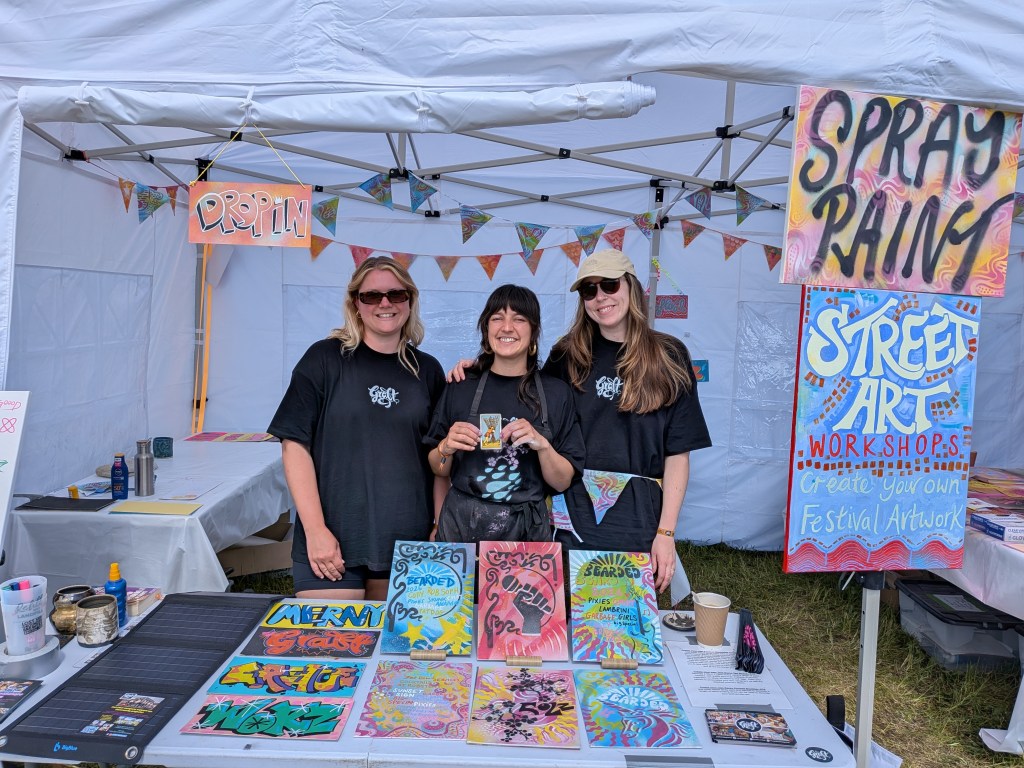 Three street art workshop organisers pose at the Spray Paint Street Art stall at Bearded Theory Festival 2026, with one holding a tarot card for Tarot Interviews.