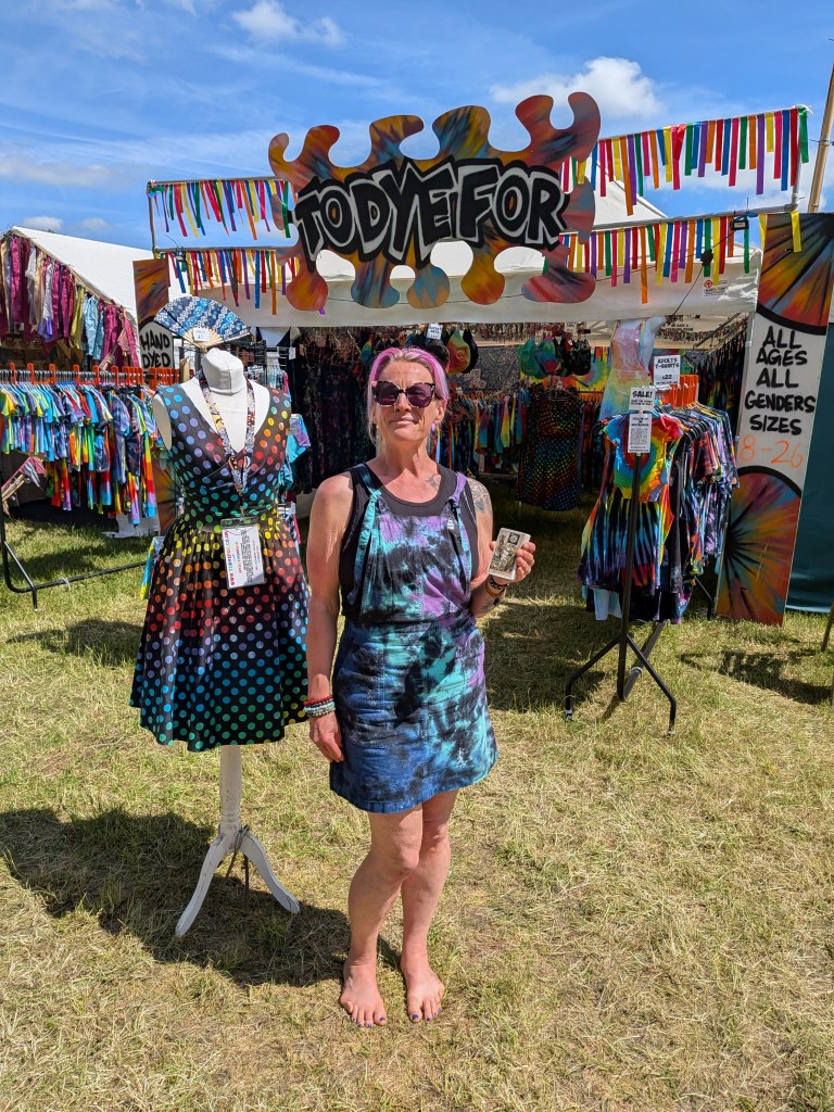 A To Dye For clothing trader poses barefoot outside a colourful festival clothing stall at Bearded Theory Festival 2026, holding a tarot card.
