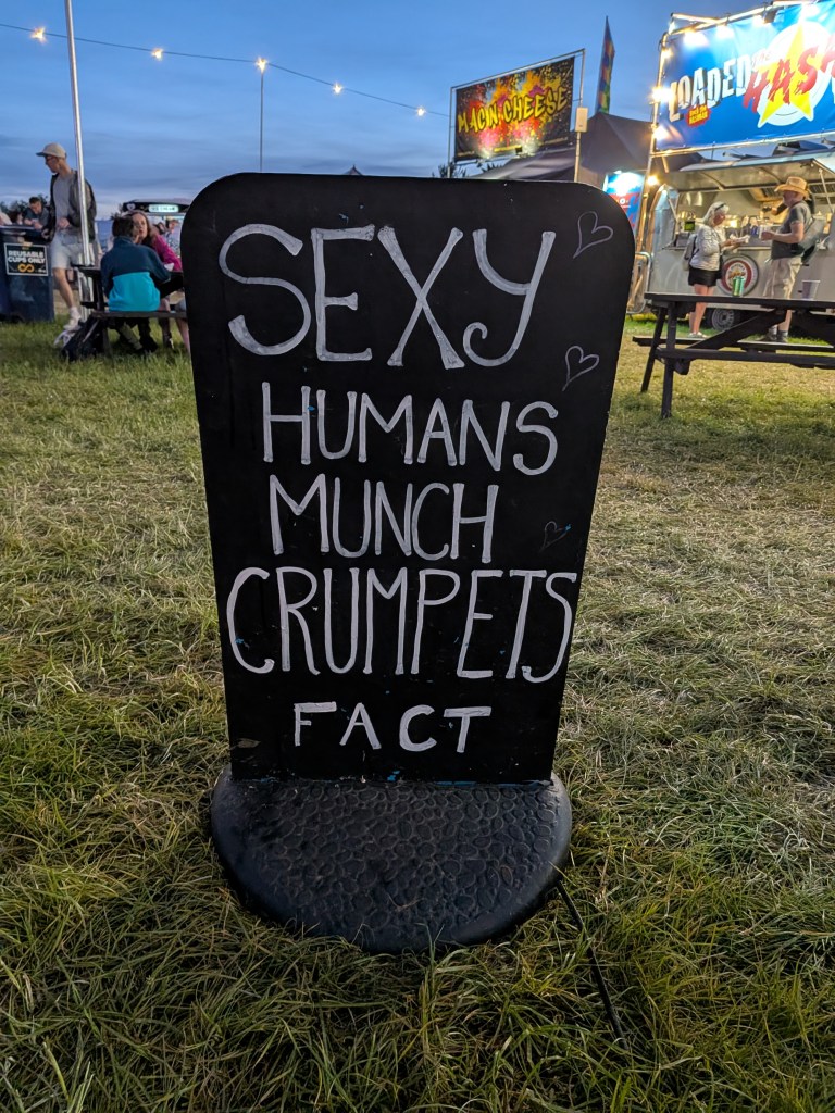 A blackboard food sign at Bearded Theory Festival 2026 reads “Sexy Humans Munch Crumpets Fact,” with food stalls and picnic tables in the background.