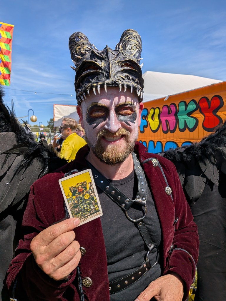 A costumed festival-goer with a horned mask, black wings and face paint holds the Five of Pentacles tarot card at Bearded Theory Festival 2026.