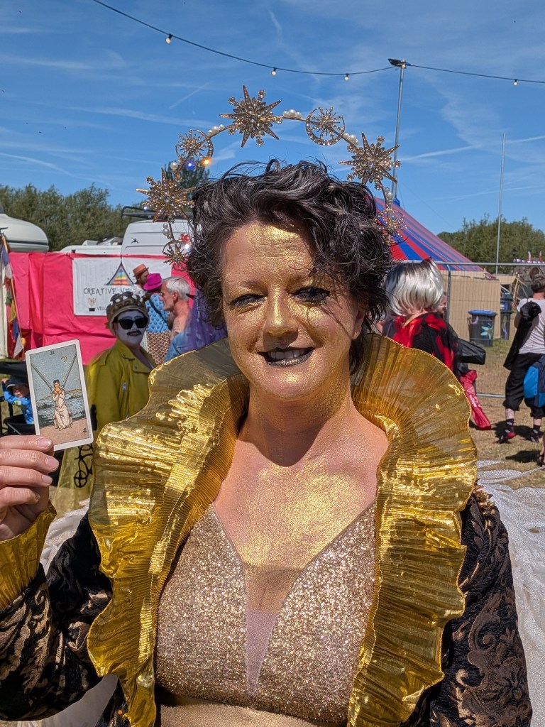 A gold-painted performer in a sparkling costume and ornate headpiece holds the Two of Swords tarot card at Bearded Theory Festival 2026.