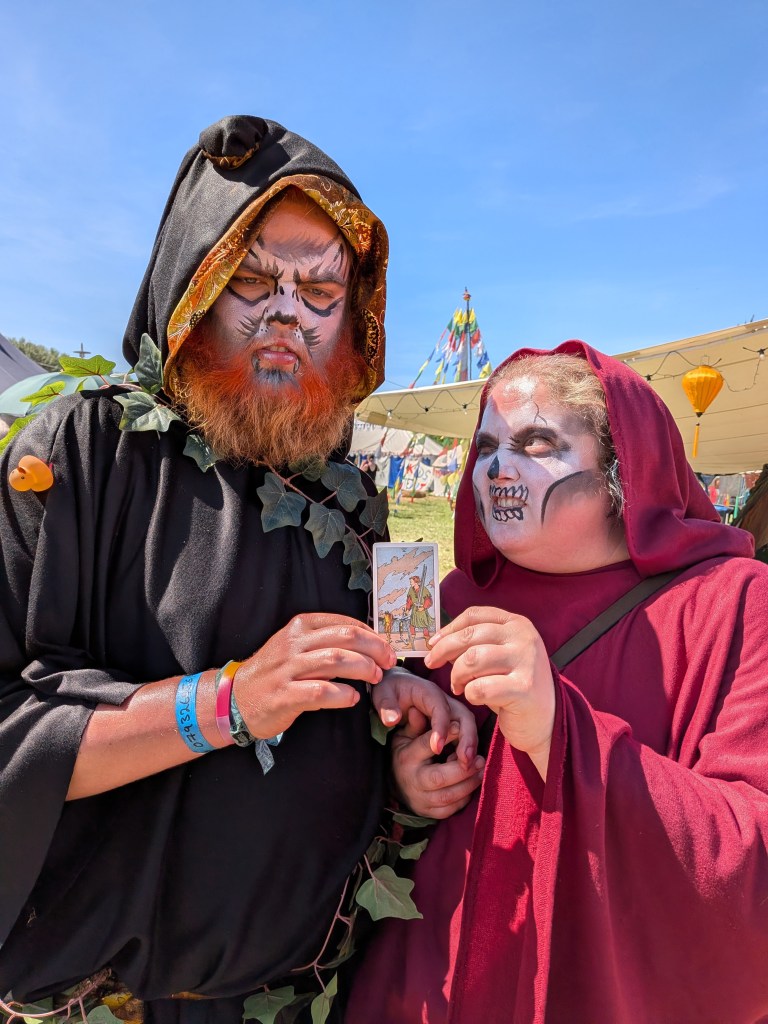 Two costumed festival-goers in hooded robes and face paint hold the Five of Swords tarot card at Bearded Theory Festival 2026.
