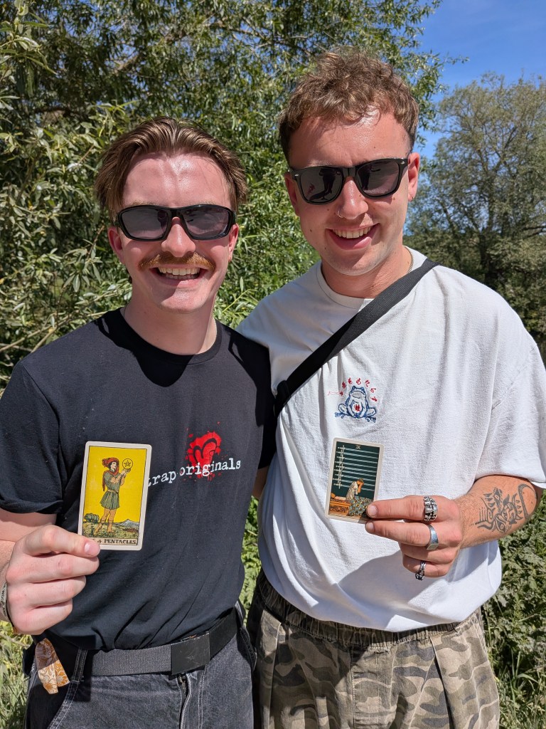 Two members of GANS smile for Tarot Interviews at Bearded Theory Festival 2026, each holding a Rider-Waite-Smith tarot card.