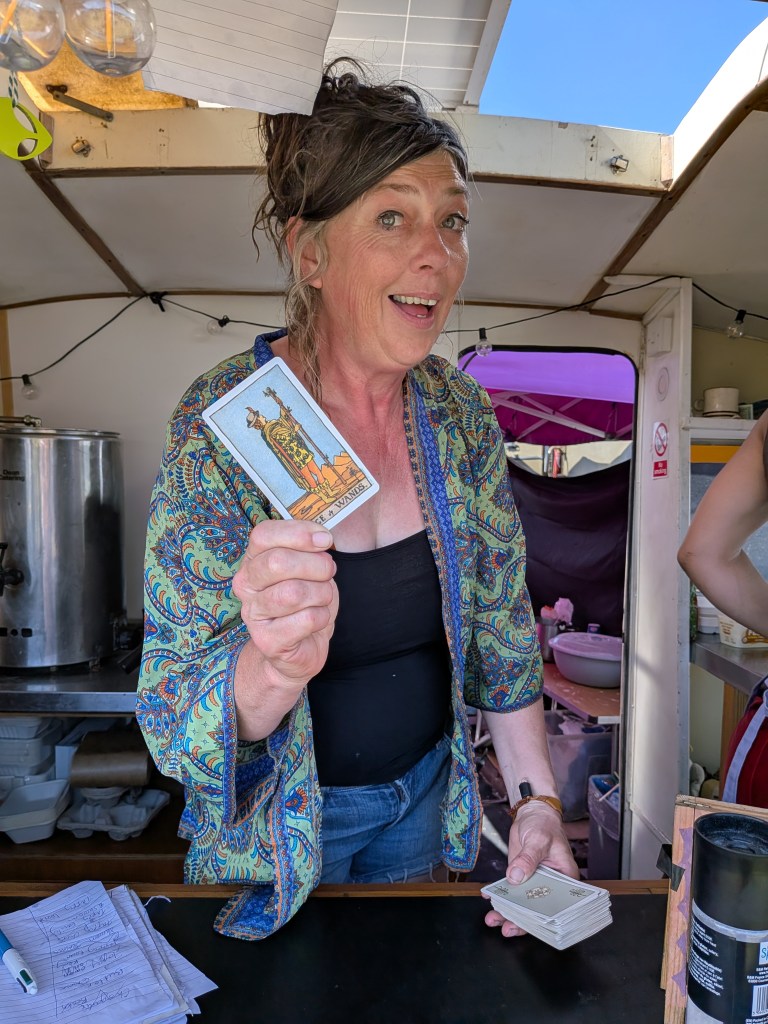 A trader at Bearded Theory Festival 2026 holds a tarot card inside a stall, with a deck and handwritten notes visible on the counter.