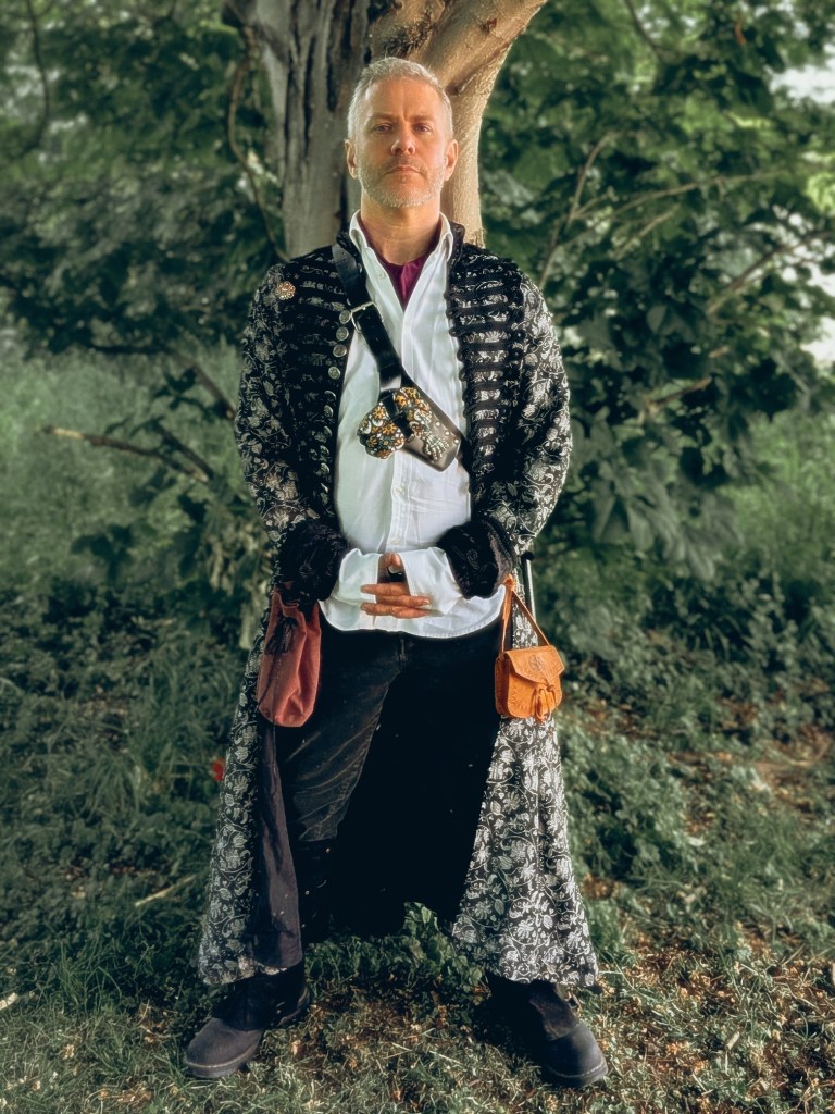 Finbarre Snarey stands beneath a tree at Wyldwood Renaissance Festival, wearing a black-and-silver embroidered coat, white shirt, belt pouch and satchel in a woodland setting.