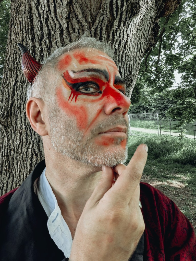 Close-up portrait of Finbarre Snarey at Wyldwood Renaissance Festival, wearing red and black devil-style face paint, a horn and renaissance costume beside a large tree.