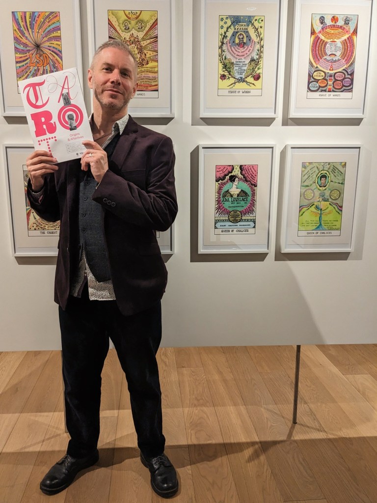 Finbarre Snarey holding a tarot-themed book at the Warburg Institute tarot event in 2025, standing in front of framed tarot-inspired artworks.