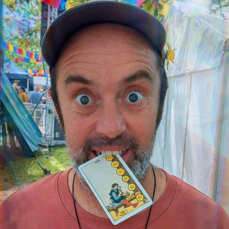 Close-up of a festival-goer playfully biting the Eight of Pentacles tarot card beneath colourful bunting and bright sunlight.