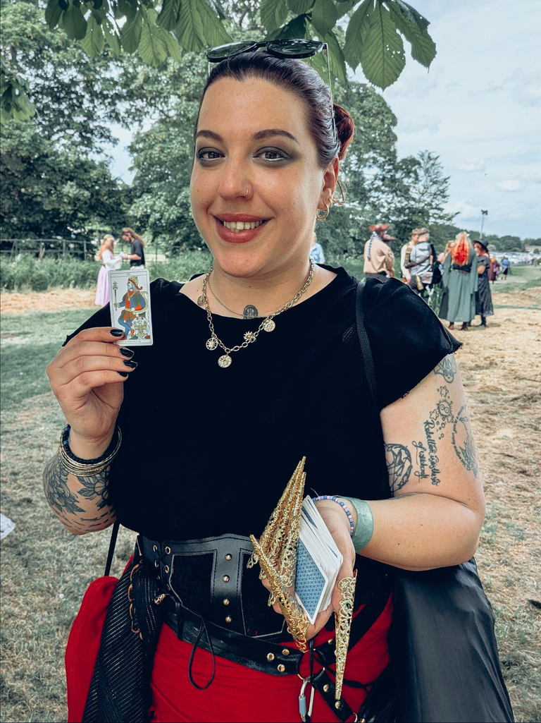 Festival-goer in a black and red outfit holding a tarot card and deck beneath leafy trees.