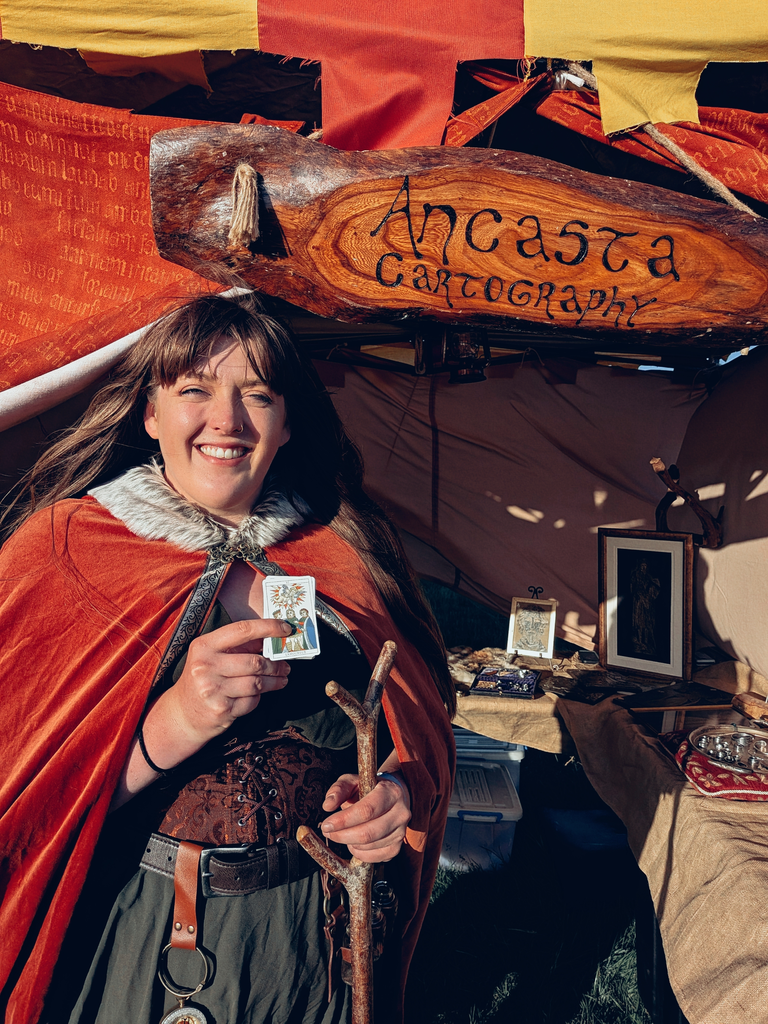 Smiling cartography trader in an orange cloak holding a tarot card outside the Ancasta Cartography stall.