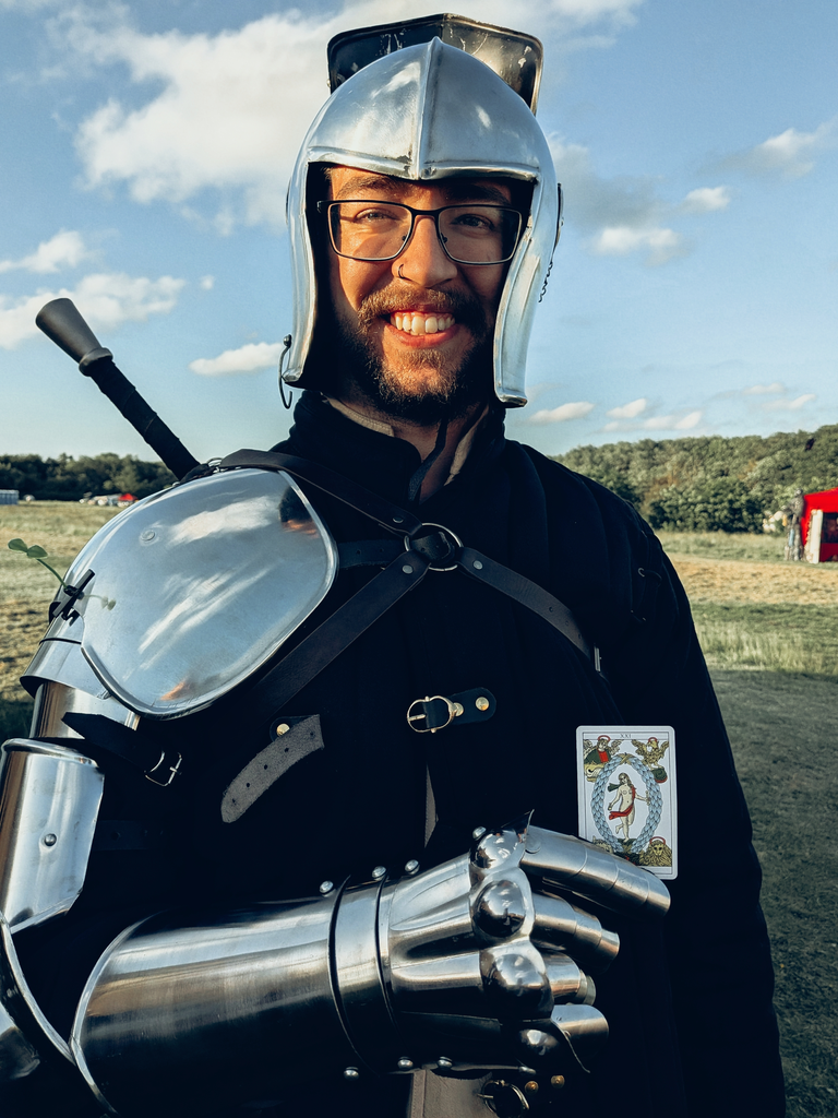 Armoured living-history performer wearing a helmet and metal gauntlet with The World tarot card tucked into his costume.