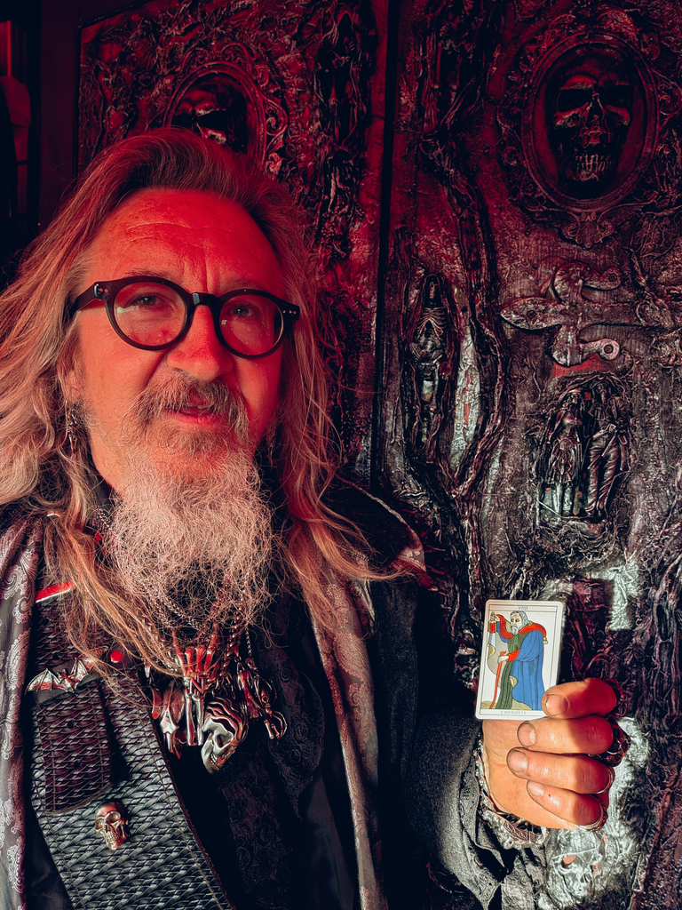 Gothic-styled festival trader holding The Hermit tarot card beside dark skull artwork in red lighting