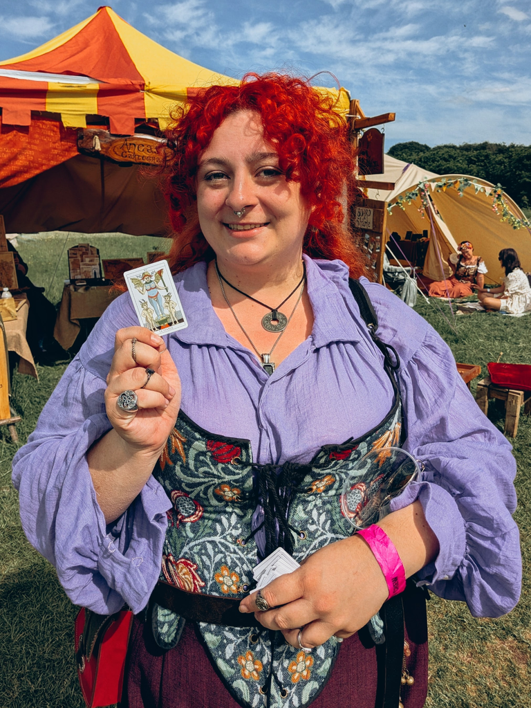 Red-haired festival-goer in a purple blouse holding a tarot card near colourful medieval-style tents.