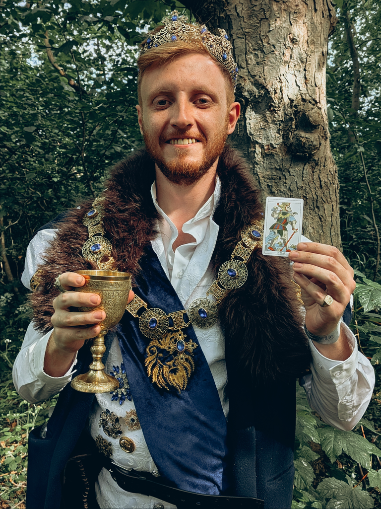 Costumed festival-goer dressed as a king holding a goblet and The Fool tarot card in woodland.