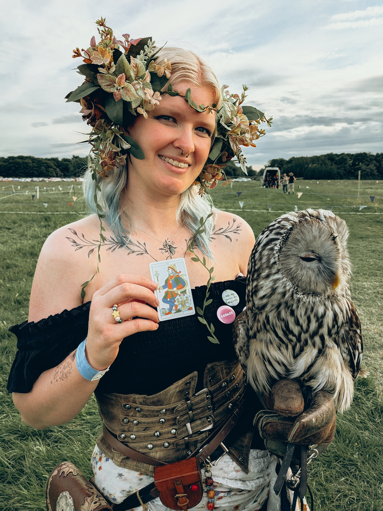 Festival-goer wearing a leafy flower crown holding a tarot card while carrying a grey owl on a glove