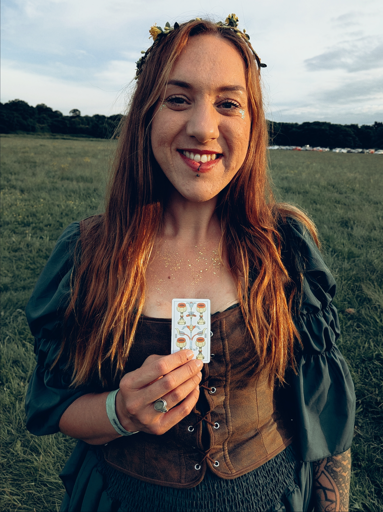 Smiling festival-goer with glitter makeup holding a tarot card in an open field at golden hour.