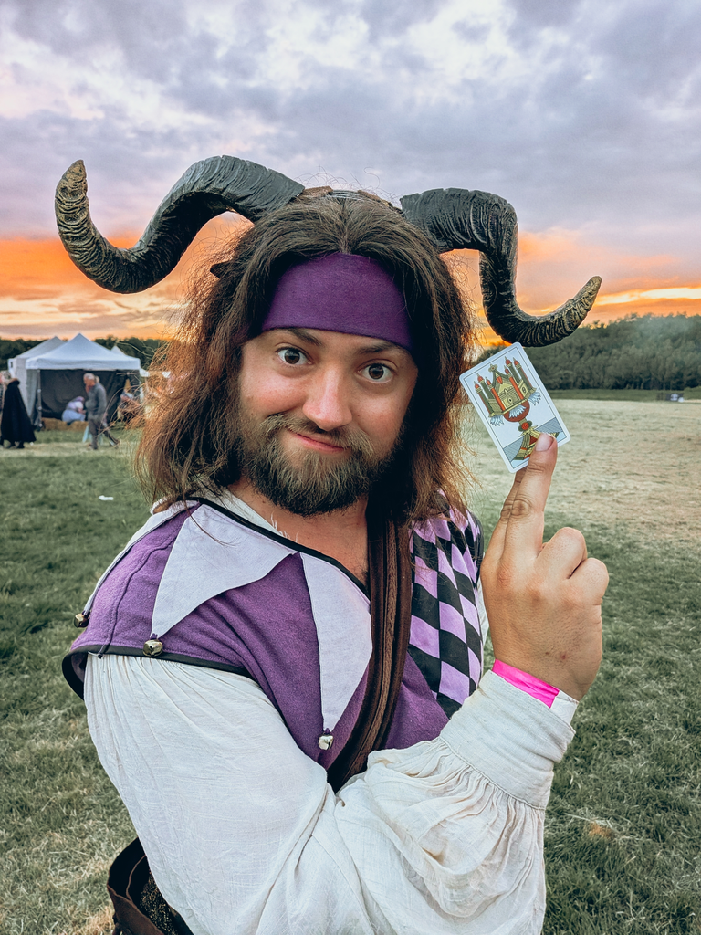 Horned festival performer in purple costume holding an upside-down tarot card against a sunset sky.