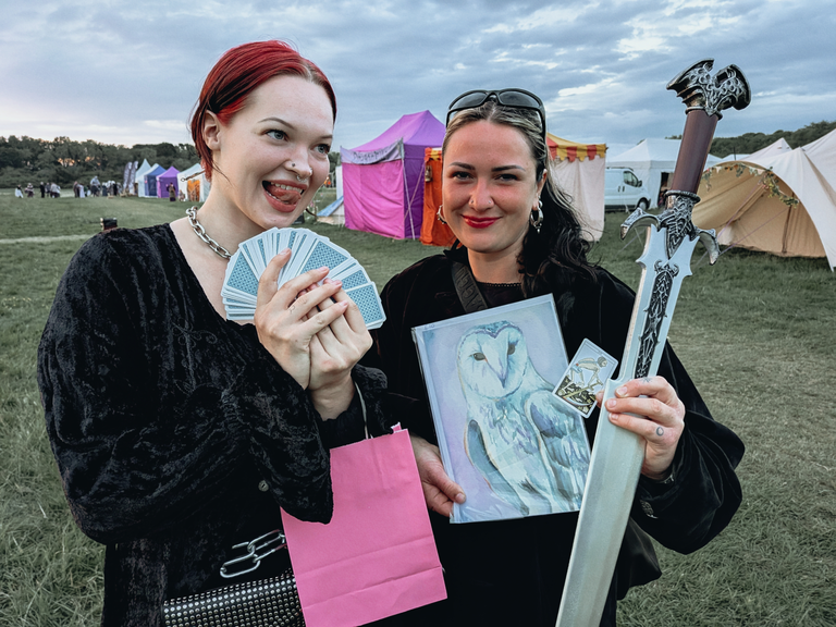 Two festival-goers in dark fantasy outfits posing with tarot cards, an owl print and a large silver sword.