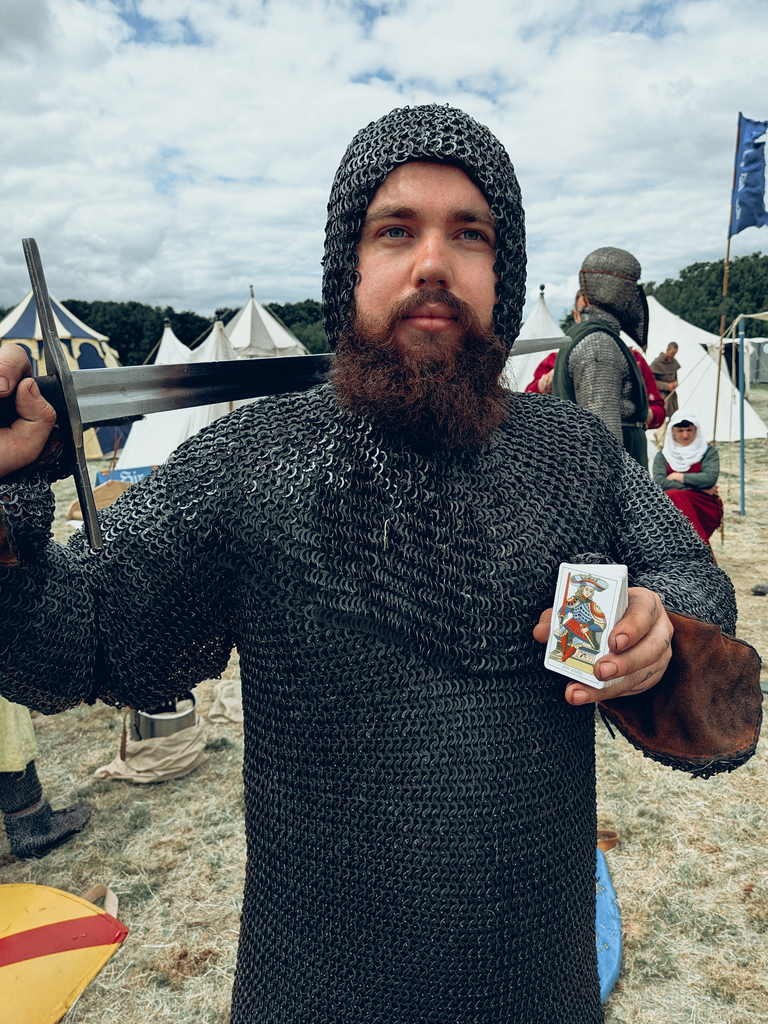 Chainmail-clad living-history performer holding a sword and tarot deck at a medieval encampment.