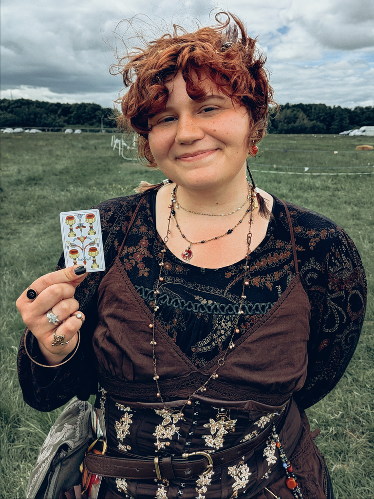 Festival-goer in a brown embroidered outfit holding a tarot card in a grassy field.