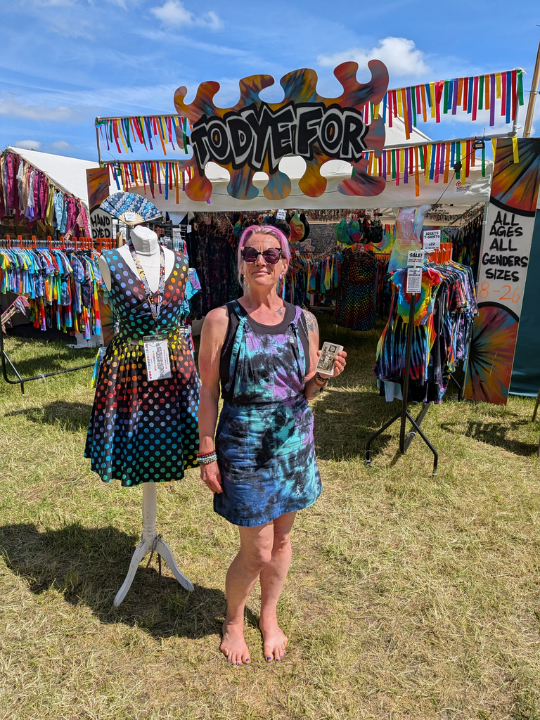 Tie-dye clothing trader standing barefoot outside a bright festival stall while holding a tarot card.