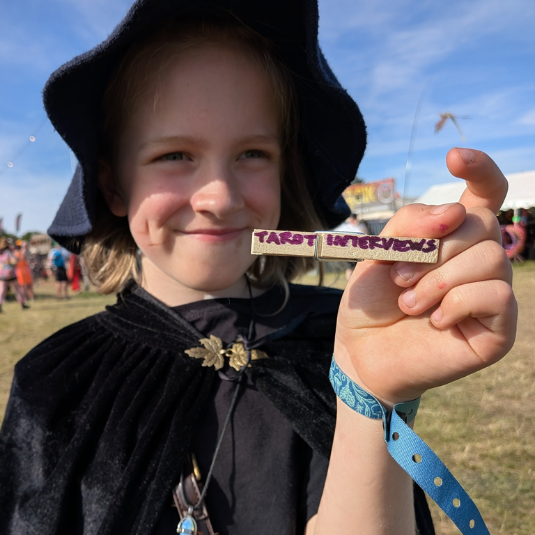 Young festival-goer in a black cloak and hat holding a small peg labelled “Tarot Interviews.”