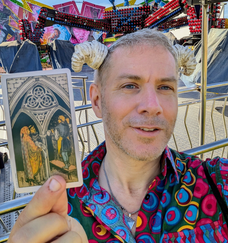 Tarot Interviews host holding the Three of Pentacles card beside a fairground ride on a sunny festival day.