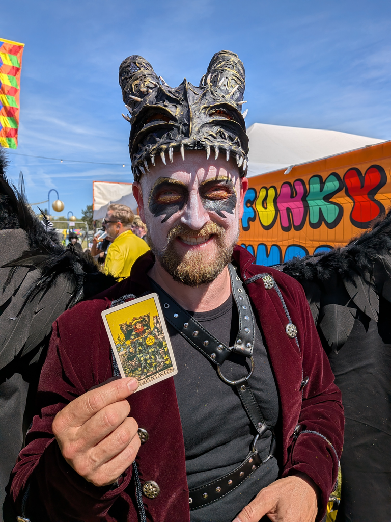 Costumed festival performer with black wings and a horned mask holding the King of Pentacles tarot card.