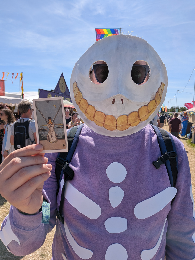 Festival-goer in a large skull costume holding the Two of Swords tarot card in front of colourful tents and flags.