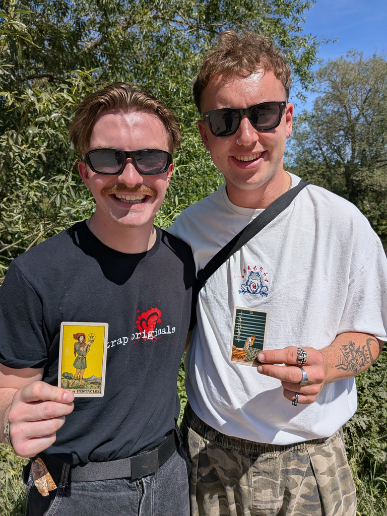 Two smiling musicians holding tarot cards outdoors at a sunny festival.