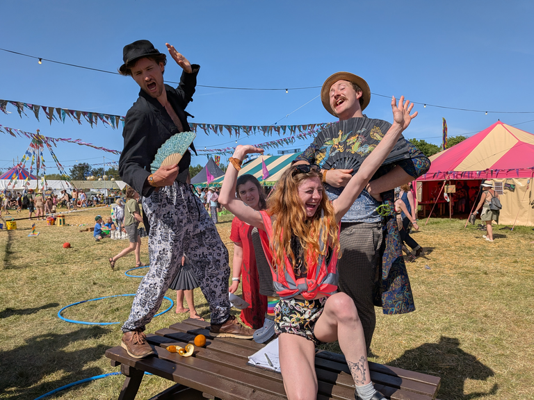 Three members of staff dance on a table at Bearded Theory in the sunshine.