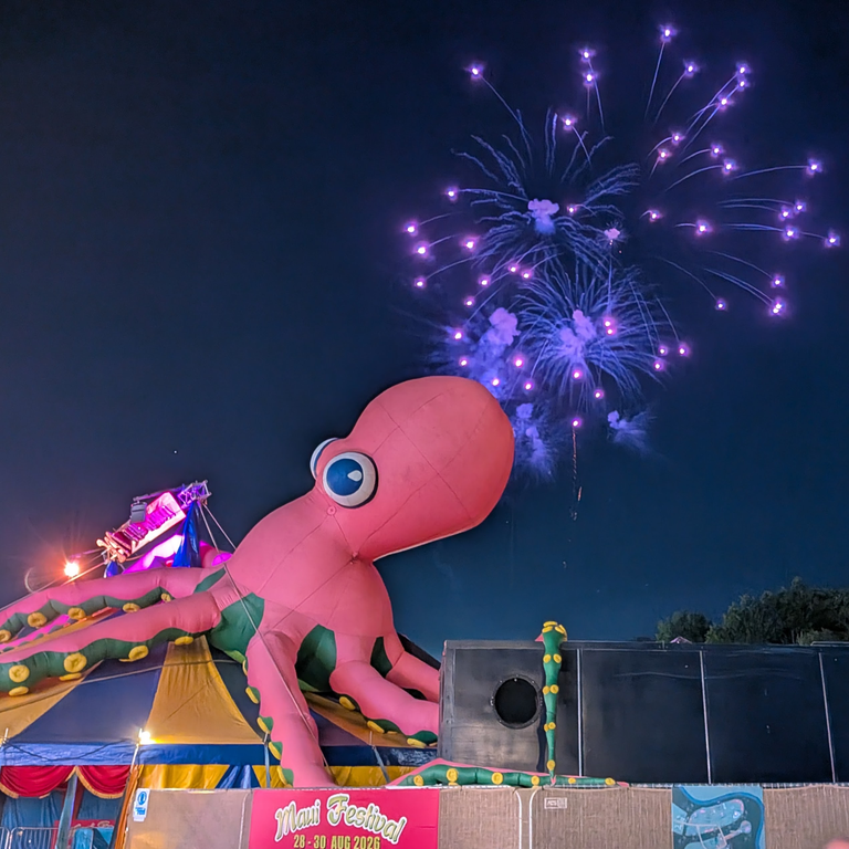 Night-time festival scene with a giant pink inflatable octopus and purple fireworks overhead.