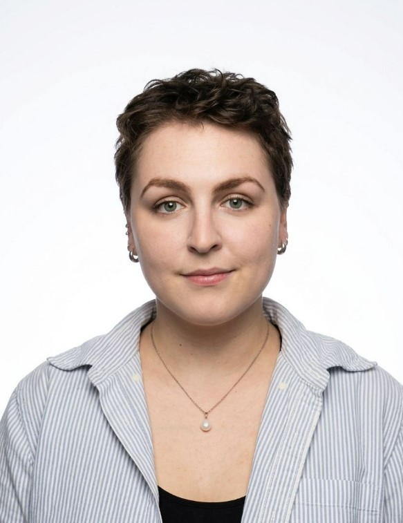 A head and shoulders photograph of a person with light skin and short, dark curly hair, looking directly at the camera with a subtle smile. They are wearing a light blue and white striped button-down shirt over a black top and a simple necklace with a small pearl pendant. They have visible piercings in their ears, including two small hoops. The background is a solid,