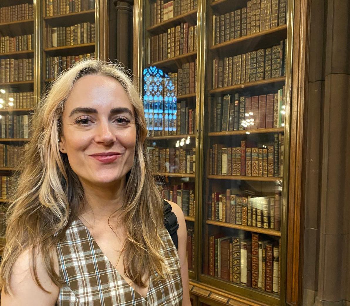 Writer Madelaine Gnewski smiles at the camera in front of tall, glass-fronted wooden bookcases filled with antique books. She has long blonde hair and wears a brown-and-cream checked sleeveless top.