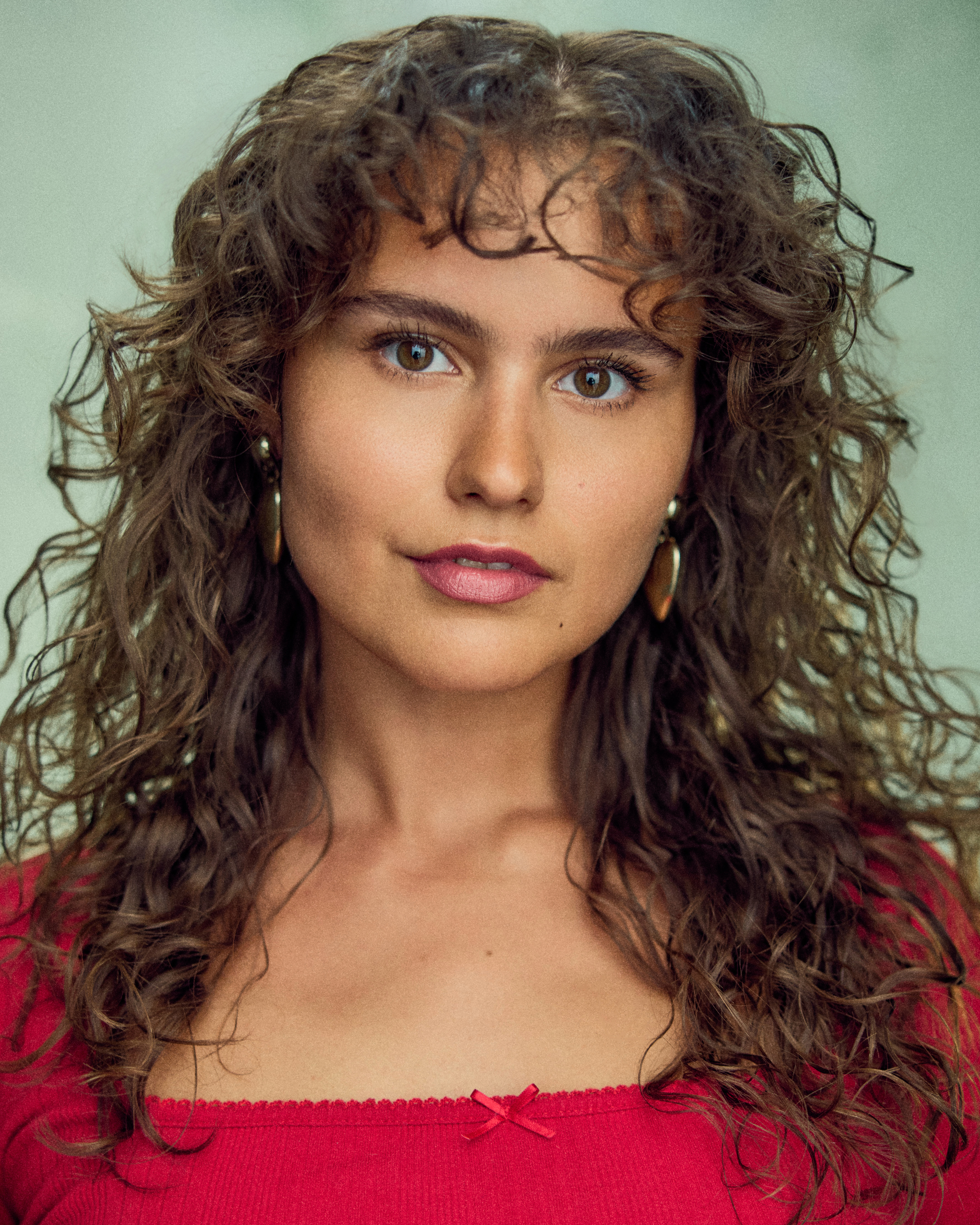 Close-up portrait of Boo Miller looking directly at the camera, with long curly brown hair, gold hoop earrings and a red top against a pale green background.