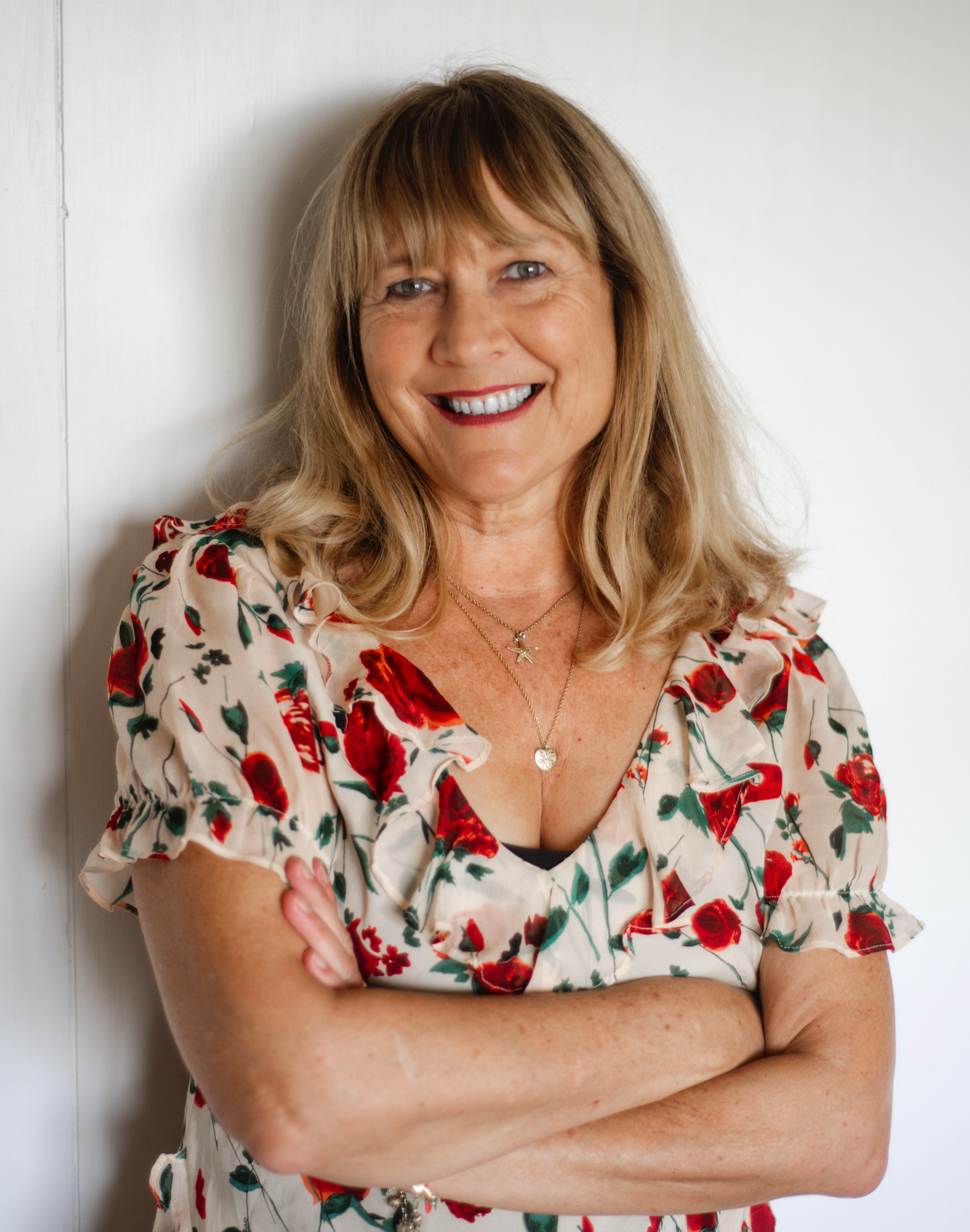Jill Dawson smiling at the camera, wearing a floral blouse and standing against a white wall.