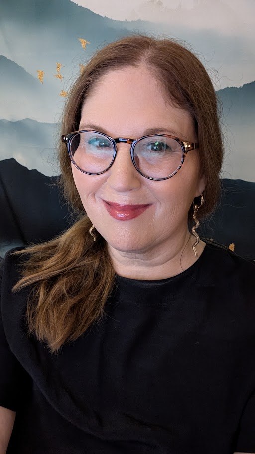 Portrait of a smiling woman with long auburn hair, round dark-framed glasses and a black top, seated against a softly coloured blue-green background.